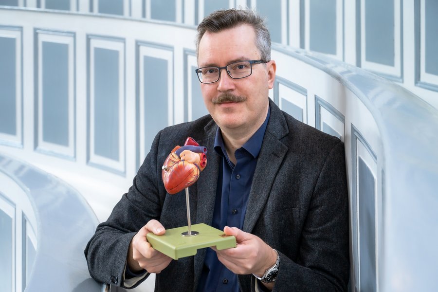 Ein Mann mit Brille, Schnauzbart und dunklem Anzug sitzt in historischen Hörsaal der halleschen Anatomie. Er hält mit beiden Händen ein Modell eines menschlichen Herzens in die Kamera.  Ein Mann mit Brille, Schnauzbart und dunklem Anzug sitzt in historischen Hörsaal der halleschen Anatomie. Er hält mit beiden Händen ein Modell eines menschlichen Herzens in die Kamera.