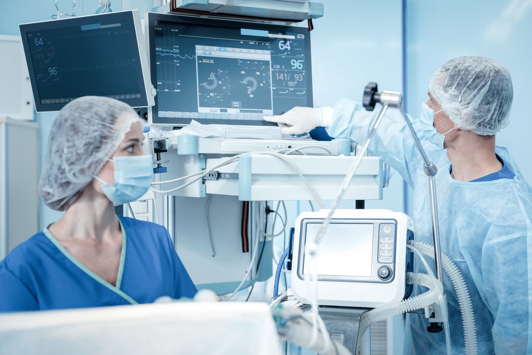 Medizinisches Personal im OP. Ein Mann zeigt auf einem Monitor auf Vitalwerte. Eine Frau schaut. Beide sind im OP-Kleidung mit Maske und Haube.