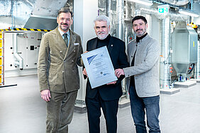 Drei Männer in Anzügen halten gemeinsam ein Zertifikat. Im Hintergrund ist ein Technikraum zu sehen.