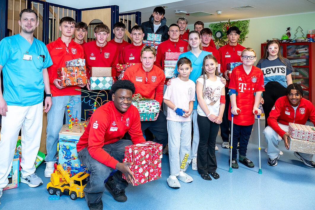 Spieler der U19-Mannschaft des HFC posieren mit Mitarbeitenden der Kinderstation des UKH und Patient:innen für ein Gruppenbild. Einige halten Geschenke in den Händen.