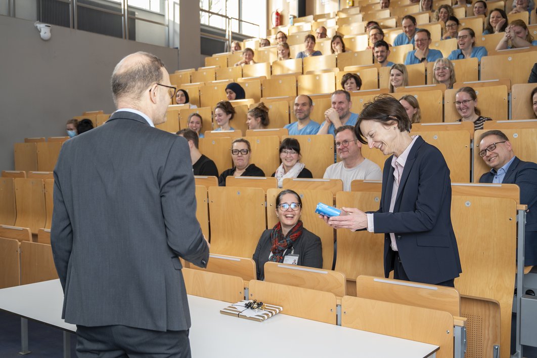 Ein Mann im Anzug steht mit dem Rücken zur Kamera vor einem Hörsaal voller Menschen. In der ersten Sitzreihe steht Simone Hettmer, die sich über ein kleines blaues Päckchen freut. Viele Menschen im Raum lachen.