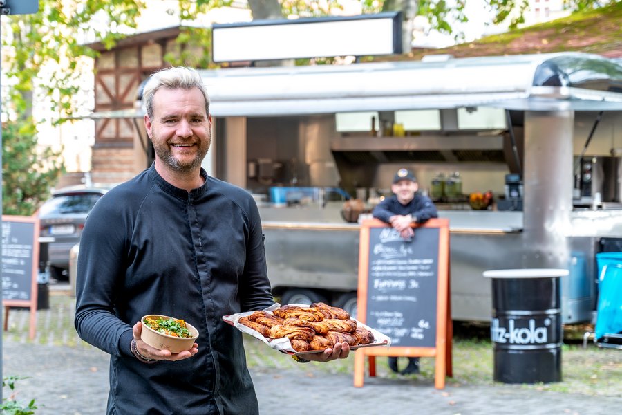 Ein Koch steht vor einem Foodtruck und hält Speisen in den Händen. Im Hintergrund lehnt eine weitere Person an einem Schild. Dahinter befindet sich der Foodtruck.  Ein Koch steht vor einem Foodtruck und hält Speisen in den Händen. Im Hintergrund lehnt eine weitere Person an einem Schild. Dahinter befindet sich der Foodtruck.