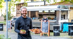 Ein Koch steht vor einem Foodtruck und hält Speisen in den Händen. Im Hintergrund lehnt eine weitere Person an einem Schild. Dahinter befindet sich der Foodtruck.