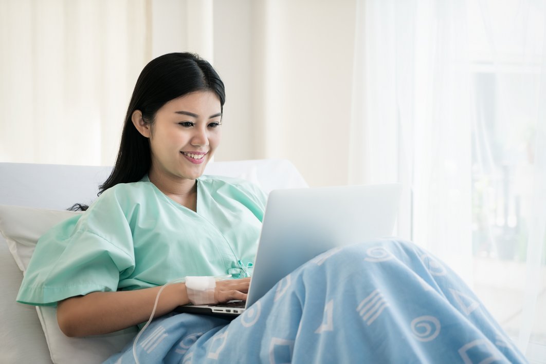 Frau sitzt im Krankenbett mit angewinkelten Beinen und arbeitet am Laptop