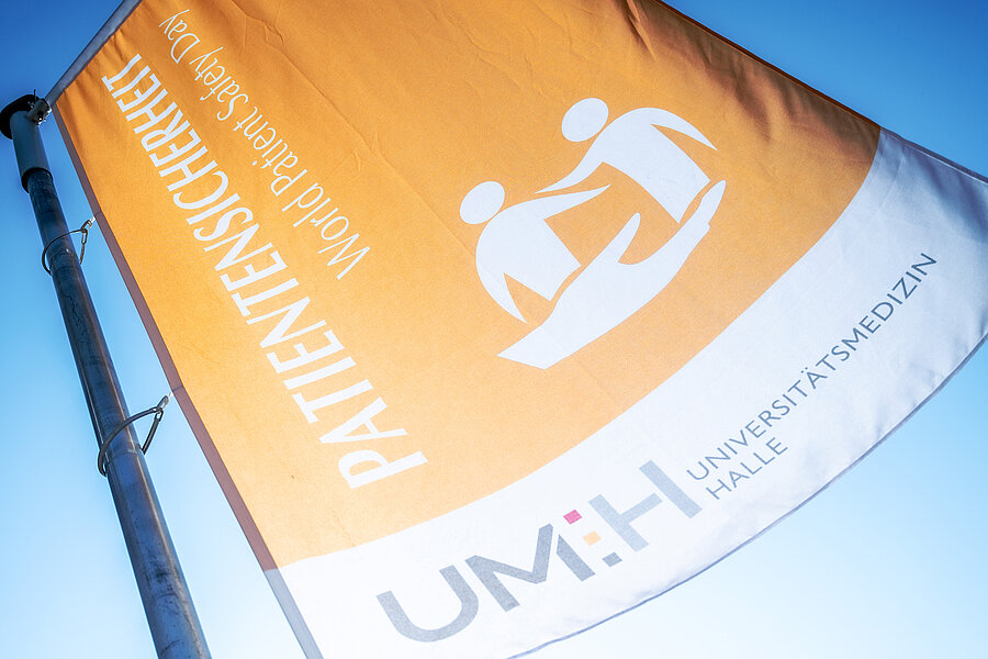 Nahaufnahme einer Fahne, die an einem Fahnenmast weht. Auf der linken Seite der Fahne steht „Patientensicherheit“ und „World Patient Safety Day“. Rechts daneben befindet sich das Logo des Welttags der Patientensicherheit (eine Hand und zwei Personen, die je einen Arm zueinander ausstrecken). Unten befindet sich das Logo der Universitätsmedizin Halle. Nahaufnahme einer Fahne, die an einem Fahnenmast weht. Auf der linken Seite der Fahne steht „Patientensicherheit“ und „World Patient Safety Day“. Rechts daneben befindet sich das Logo des Welttags der Patientensicherheit (eine Hand und zwei Personen, die je einen Arm zueinander ausstrecken). Unten befindet sich das Logo der Universitätsmedizin Halle.
