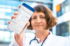 Eine Person in medizinischer Arbeitskleidung hält ein Smartphone in die Kamera. Dieses zeigt die geöffnete Telekasper-App.